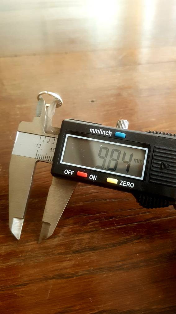 A digital caliper measures a small 18-gauge metal object, displaying a measurement of 9.04 millimeters on its screen. The caliper is held over a wooden surface, with its "on" button illuminated red, perfect for ensuring precision while crafting the Kaya Movement AYM Seamless Septum Ring in Gold for body jewelry.